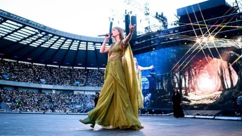 Getty Images A woman holding a microphone and wearing a greed dress walks across a large stage with thousands of concert goers visible in the stands of a stadium behind her