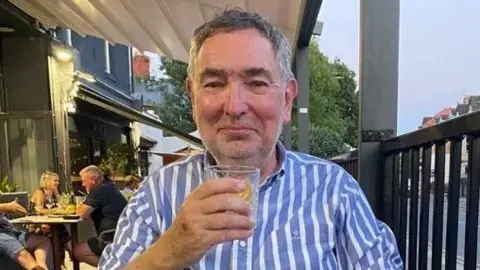 Aled, a man with black and grey hair wearing a blue and white striped shirt. He is smiling at the camera while holding a glass with a drink inside. He is sat in outdoor area of a bar/restaurant.