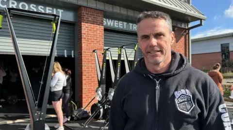 Gym owner Adam Shepherd looking at the camera outside his gym. There is some equipment outside the gym, which is in an industrial unit, and people are working out behind him.