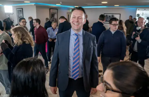 PA Media Russell Findlay in a suit and striped tie stands in the middle of a busy indoor event, surrounded by groups of people talking, taking photos and moving around the room, with bright lighting and framed photographs on the walls