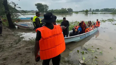आझादहे मोशीरी/बीबीसी बचावकर्ते लोकांना पूरग्रस्त नदीच्या काठावर एका छोट्या बोटीवर मदत करतात