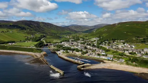 An aerial view of the coastal community of Helmsdale in the Highlands. Helmsdale is at the mouth of a river, and has a harbour and rows of white-walled houses. Green hills rise behind the village.