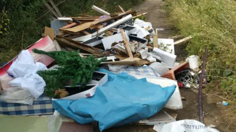 A pile of fly-tipping in a county lane. Pieces of wood, electricals, mattresses and Christmas trees are visible.