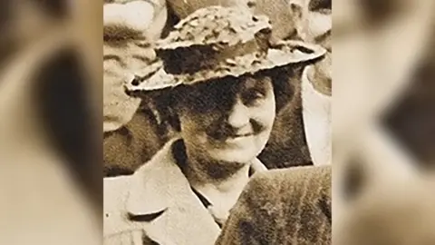 Avon and Somerset Police Louisa Dunne in the 1940s, stood among a crowd in a black-and-white photo. She is wearing a formal coat and brimmed hat, and is smiling.