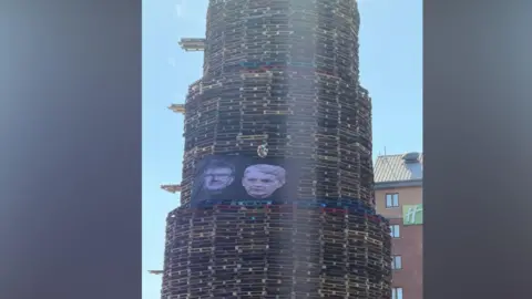 A towering bonfire built from wooden pallets stands on a city street on a sunny day.  On the side of the bonfire there is a large black banner showing photos of the faces of Jeffrey Donaldson and his wife Eleanor.  There is a red brick hotel in the background. 