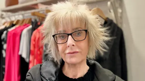 BBC A close up shot of a woman with short blonde hair. She has black, square-framed spectacles on her face and she is looking directly down at the camera. She is wearing a black coat over a black top and behind her is a clothing rail, on which dresses and tops are hanging from beige-coloured wooden hangers.