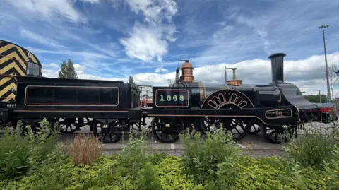 A black steam engine with gold numbers and designs on the side is coupled to another small rail vehicle behind it. It is on the back of a vehicle that is transporting it. There is grass and bushes in the foreground and a blue sky.