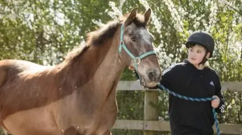 Family Handout A boy is leading a horse along a fenced area. The boy is wearing a black helmet and is looking at the horse, which is brown.