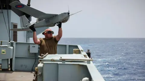 LPhot Matt Bradley A man wearing a khaki T-shirt and goggles lifts a Puma drone over his head to launch it from a ship with the sea seen beyond 