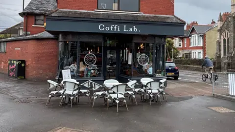 A Coffi Lab cafe on the corner of a street. The building is red brick with grey signage. White chairs are outside. 