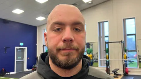 BBC / Amanda White. La foto muestra a un hombre sonriendo a la cámara. Es calvo, pero tiene barba castaña. Lleva una sudadera negra y gris, y está de pie en un gimnasio. Hay pesas a su derecha.