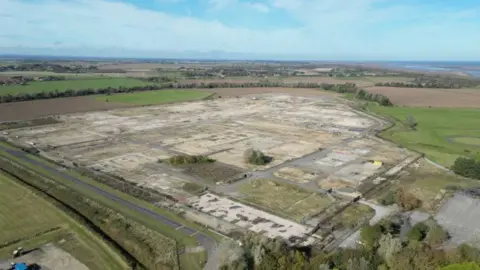 A drone view of a disused gas terminal. Little remains except roadways and hard standing. The extensive site is bordered by fields. In the distance, hedgerows and trees give way to a beach and the sea.
