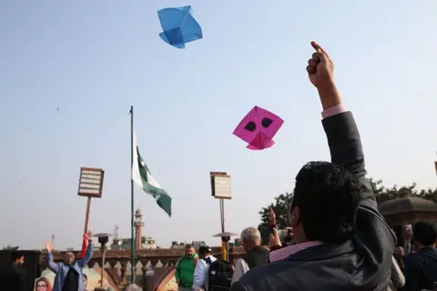 Anadolu via Getty Images A man lets off a kite during the Basant kite festival in Lahore, Pakistan, as seen from behind him with his hand in the air and the kites flying off in the blue sky.