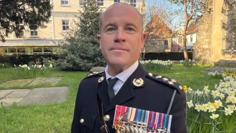 Brigadier Simon Rogers wearing a black suit with decorated epaulettes and a strip of colourful wartime medals. He is standing in the churchyard on a sunny day, surrounded by daffodils.