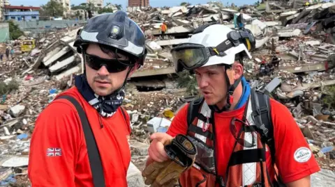 ECFRS Rob Wingar is talking to a fellow volunteer in front of a huge pile of rubble. Both are wearing orange and protective helmets.