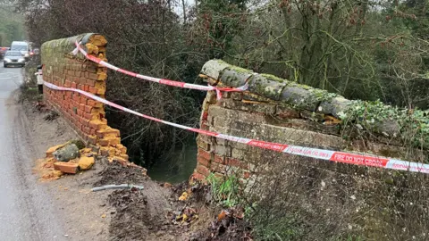 A bridge with a brick structure on Boot Street in Great Bealings, Suffolk. The bridge has been cordoned off with red and white tape across one of the walls, which has been knocked down. There is a gaping hole in the wall of the bridge.