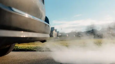 A parked car is on the road. At the back of the car there is a large amount of white and grey smoke coming from the exhaust pipe.