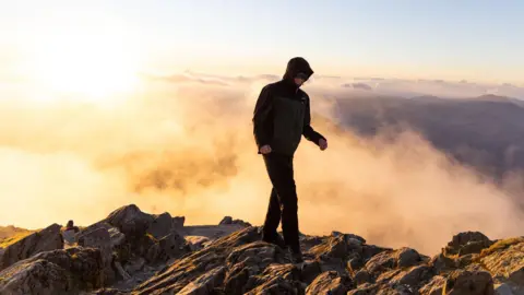 ConorHikes A man is standing on the rocky summit of a mountain wearing a coat. Behind him are cloud inversions lit up by sunrise.