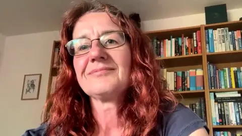 Anne Thorpe with shoulder-length curly hair wearing glasses, seated indoors. Behind her is a wooden bookshelf filled with books arranged both vertically and horizontally. 