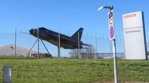 A replica of a Lightning fighter jet stands outside the main gate of the BAE Systems manufacturing site at Samlesbury, near Preston. The site is surrounded by fencing with barbed wire at the top. Outside, on a grassy bank, is a grey, vertical, rectangular block sign saying BAE Systems and Samlesbury.
