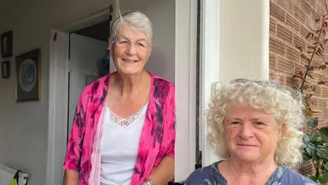 Sally Kidd (left) and Marion Parker (right). The pair are stood in a doorway with Kidd wearing a pink jacket and Parker is wearing a blue floral top.