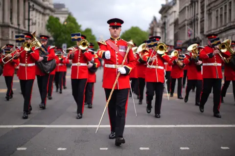 PA Media Military members in bright red uniform, carrying gold sceptres and playing brass instruments, walk down the street.