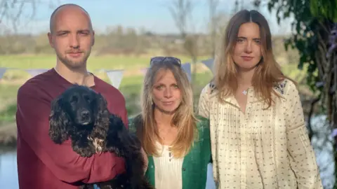 Will, Vicky and Mary Hoddinott and their dog Lola, on the riverbank close to where Alex's body was eventually found in April 2023. The sun is shining with the river and fields in the back ground.