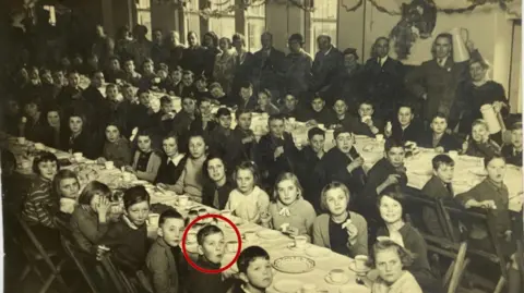 A black and white picture of children sitting at two tables in Alcombe.  They are eating food.  Adults are standing infront of windows in the background.