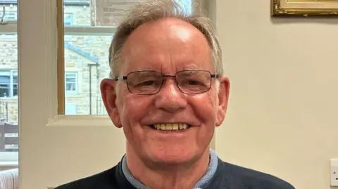 An older man with short grey hair and glasses smiles at the camera. He wears a black jumper and a blue shirt.