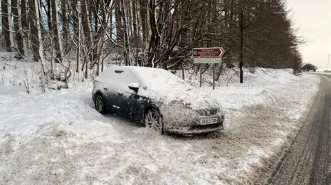 BBC A car covered in snow at the side of a rural road.