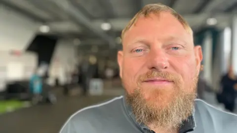 Mark King with a beard, wearing a grey top