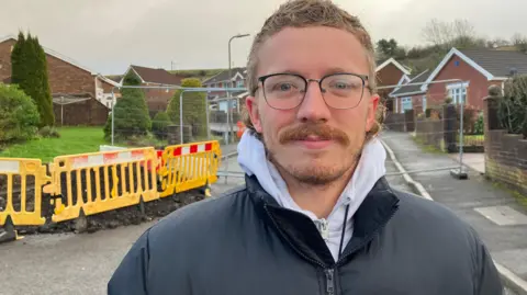 Callum Morgan stands outside the cordon. He is wearing a black winter coat with a white hooded top. He has glasses, blonde hair and a moustache and beard.