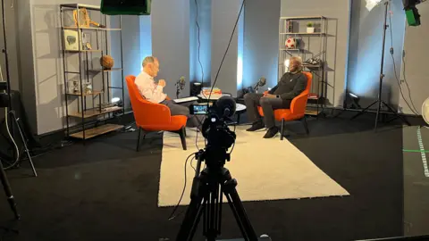 A behind the scenes shot of Jeff Brown, in pale pink shirt and grey trousers, talking to Gary Bennett, who is wearing a black jumper ad dark grey jeans. In the foreground, there is a microphone and a camera placed on a tripod. 