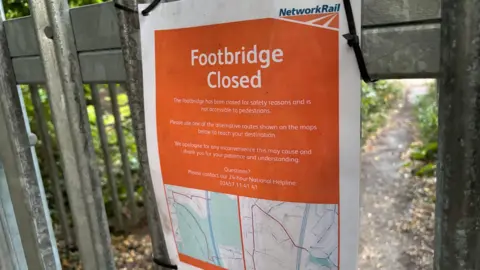 Rob Kirby/Oxhey Village Environment Group A Network Rail notice announcing the closure of a footbridge is attached to a metal gate with cable ties. The sign explains that the bridge is closed for safety reasons and provides details of alternative walking routes. Trees and a narrow path are visible in the background.