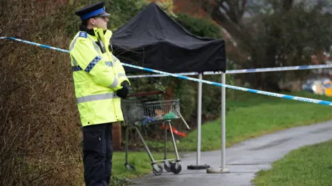 Dan Jessup A police officer stood beside the cordoned scene of an attempted murder in Eastbourne, East Sussex. A trolley can be seen in the background under a black canvas. 