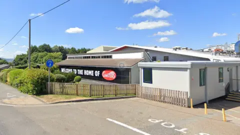 Google A pale coloured single storey industrial unit by a road with the words welcome to the home of Ginsters.