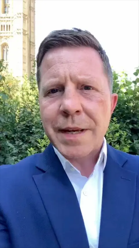 Male journalist wearing a blue suit with a white shirt, standing in front of a hedge