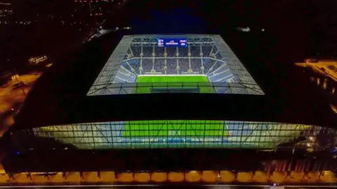 A drone view of Everton's new stadium, showing the pitch lit up at night.