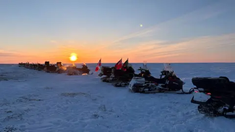Nadine Yousif/BBC The patrol snowmobiles lines up on the border between the territory of Nunavut and the province of Manitoba. The sun is seen setting across the flat snow-covered terrain.