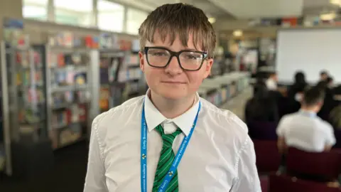 Sam Read/BBC James smiles at the camera, wearing his white school shirt, green and black striped tie and a blue lanyard saying Corby Business Academy