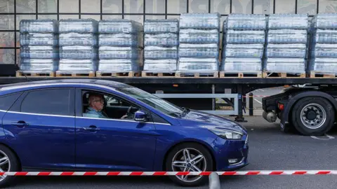 A man driving a blue car. The vehicle is stopped next to an open lorry trailer, which contains pallets of bottled water.