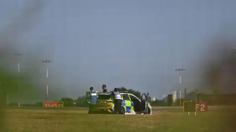 Getty Images A police car in a field. The doors are open and officers are stood around the back of the car looking to the right. They appear to be near the runway and there are signs on the ground reading F2 and F1.