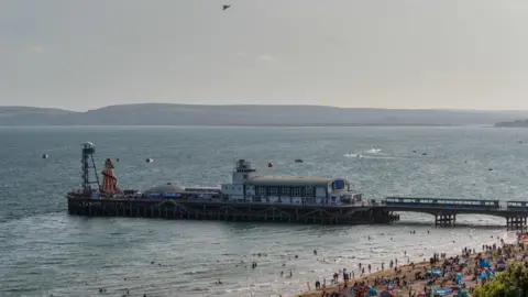 An aerial shot of Bournemouth Pier taken during the day. It comprised several buildings and structures, including a traditional helter-skelter slide. To the right, the edge of the beach is visible, with dozens of people gathered around by the sea. 