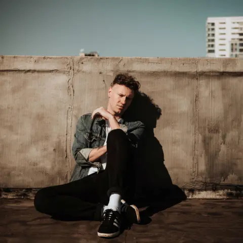 A promo shot of Callum Beattie sitting down while leaning against a wall. He is outside and there is the top of a block of flats visible. The sky is clear and blue.