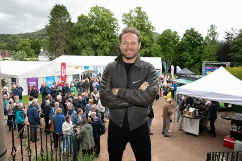 Borders Book Festival Actor Sam Heughan - a man in a black leather jacket with his arms folded - stands in front of a crowd at the Borders Book Festival