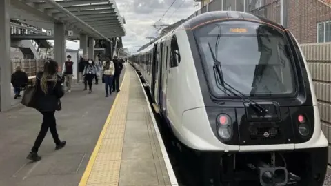 The front of an Elizabeth line train with a big glass front and white sides on the platform of an unidentified outdoor station with lots of people on the platform