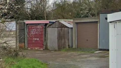 Google A row of garages with different coloured frontages stands against a backdrop of trees. The garages appear to be run down.