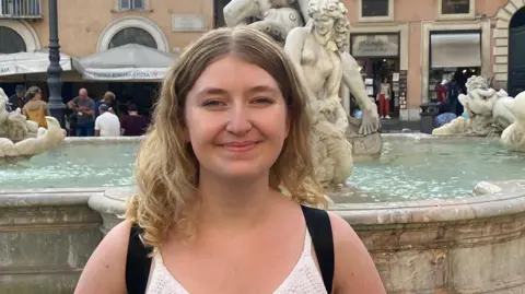 PA Media Abigail Eggleston is smiling at the camera. She is wearing a sleeveless top and has blonde shoulder-length hair. She is standing in front of a fountain in Italy.