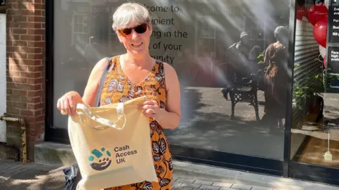 A woman with short grey hair stands outside a banking hub. She is wearing an orange dress with a brown flowered pattern. She holds a tote bag which has a printed logo reading Cash Access UK.