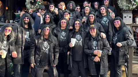A large group of people wearing black coats and trousers and t-shirts which have a picture of the face of Ozzy Osbourne on. The people are also wearing bluey-purple sunglasses, with many also wearing long dark wigs. 
